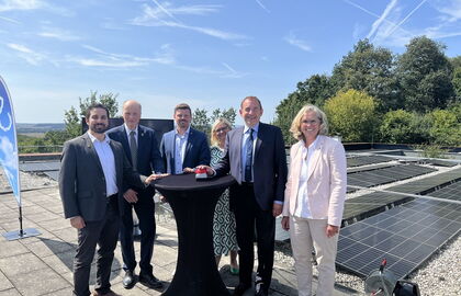 Hessens Finanzminister Professor Dr. R. Alexander Lorz steht mit 5 weiteren Personen an einen Tisch und drückt symbolisch einen Startknopf; im Hintergrund ist eine Photovoltaikanlage zu sehen